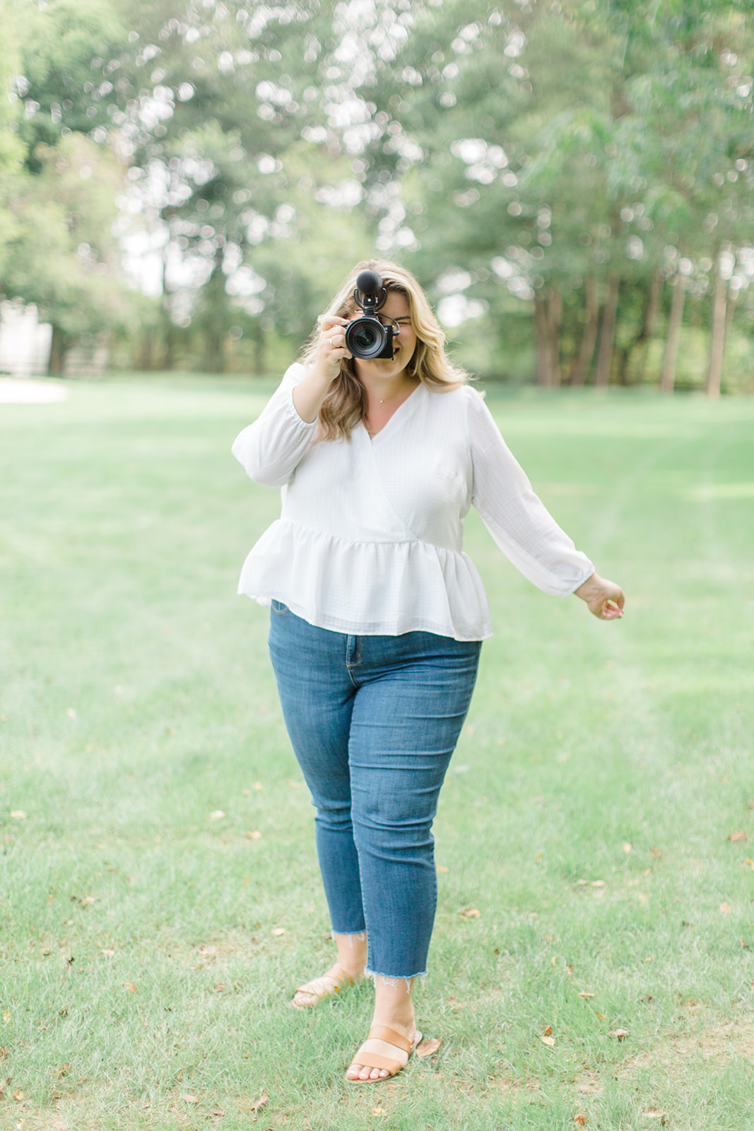 a blonde woman holding a camera to take a picture wearing a white top, straight leg jeans and sandals