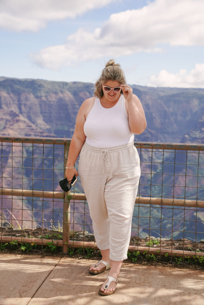 A plus size Caucasian woman is smiling at a canyon while wearing linen pants and a white tank top. 