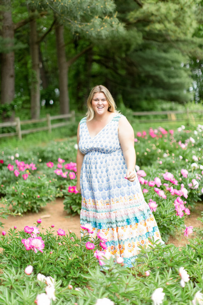 A happy Caucasian woman is posing for a picture in a garden wearing a plus-size dress from Anthropologie. 