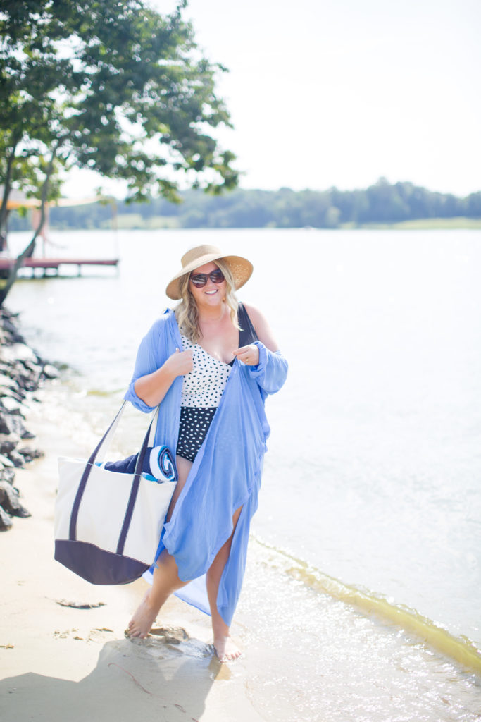 A plus size Caucasian woman walking on the beach wearing a black and white bathing suit, oversized denim shirt and straw hat, carrying a beach bag.