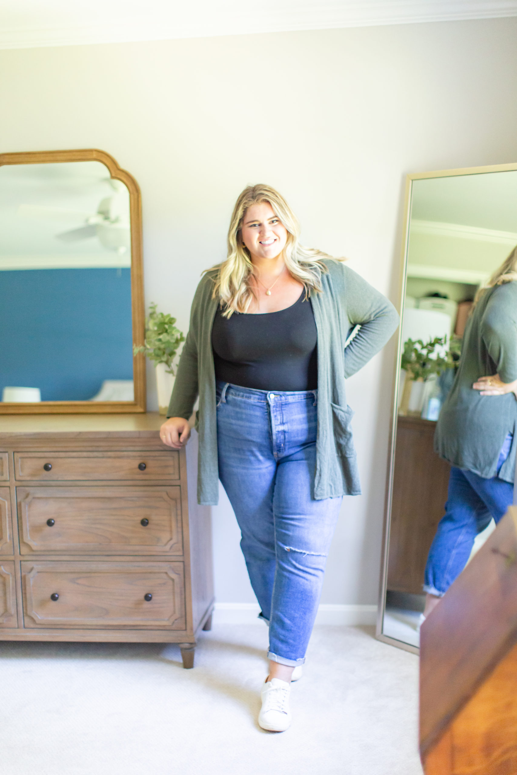 a happy Caucasian woman wearing a black tank, straight leg jeans, a green cardigan and white sneakers posing in her bedroom