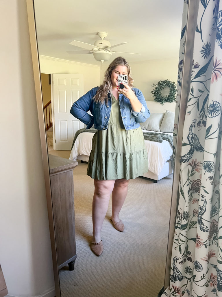 A plus-size blonde woman is taking a mirror selfie in her bedroom while modeling a plus-size dress and denim jacket. 