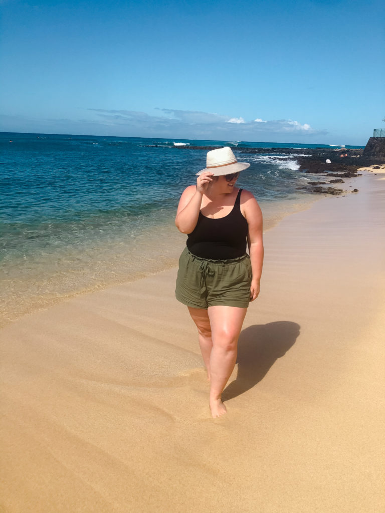 A Caucasian woman is walking on the beach wearing linen shorts and a black bathing suit. 