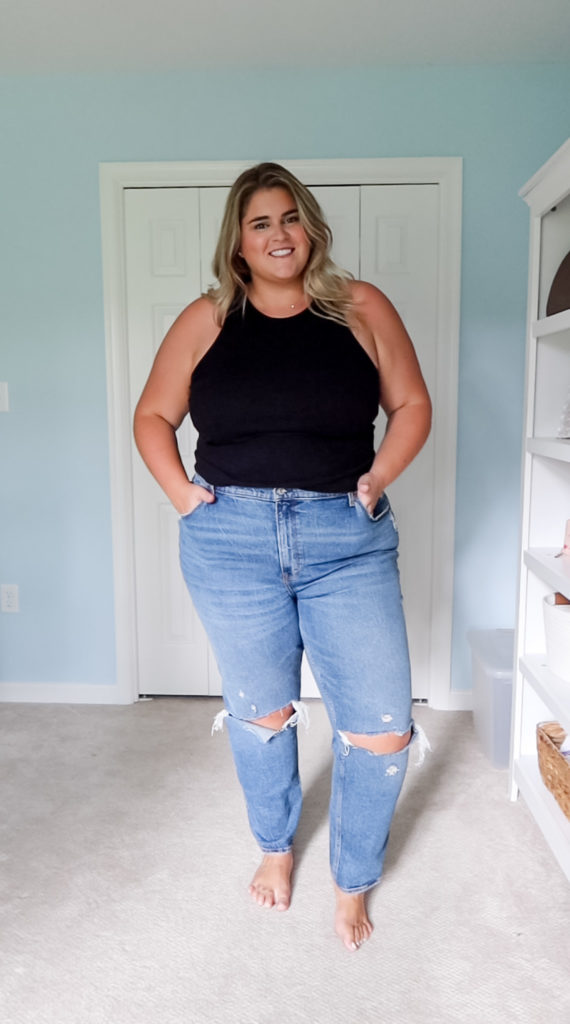 a plus size blonde woman with a fair complexion wearing plus size straight jeans and black tank top standing in her bedroom