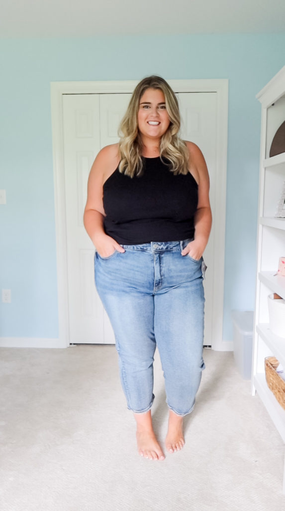 a blonde woman wearing light colored blue jeans and a black tank top posing in her bedroom