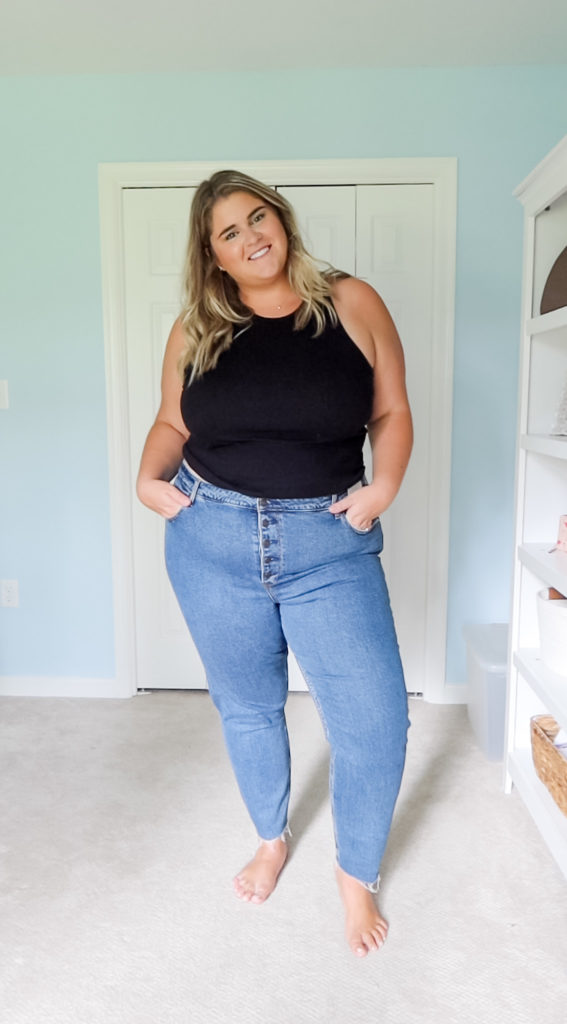 a fair-complected blonde woman wearing plus size jeans and a black tank top in her room