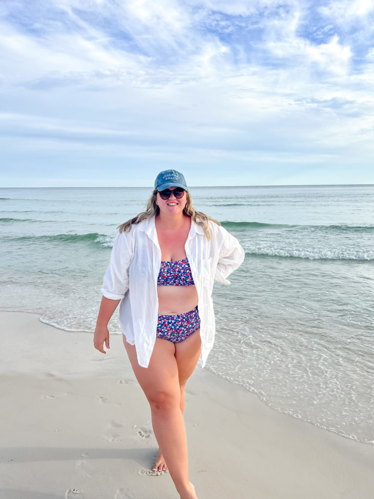 A plus size blonde woman is wearing a floral bikini, white button down and baseball cap while walking on the beach. 
