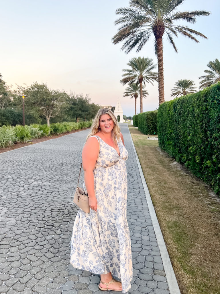 A plus size blonde woman wearing a floral maxi dress on a cobblestone path lined with palm trees. 