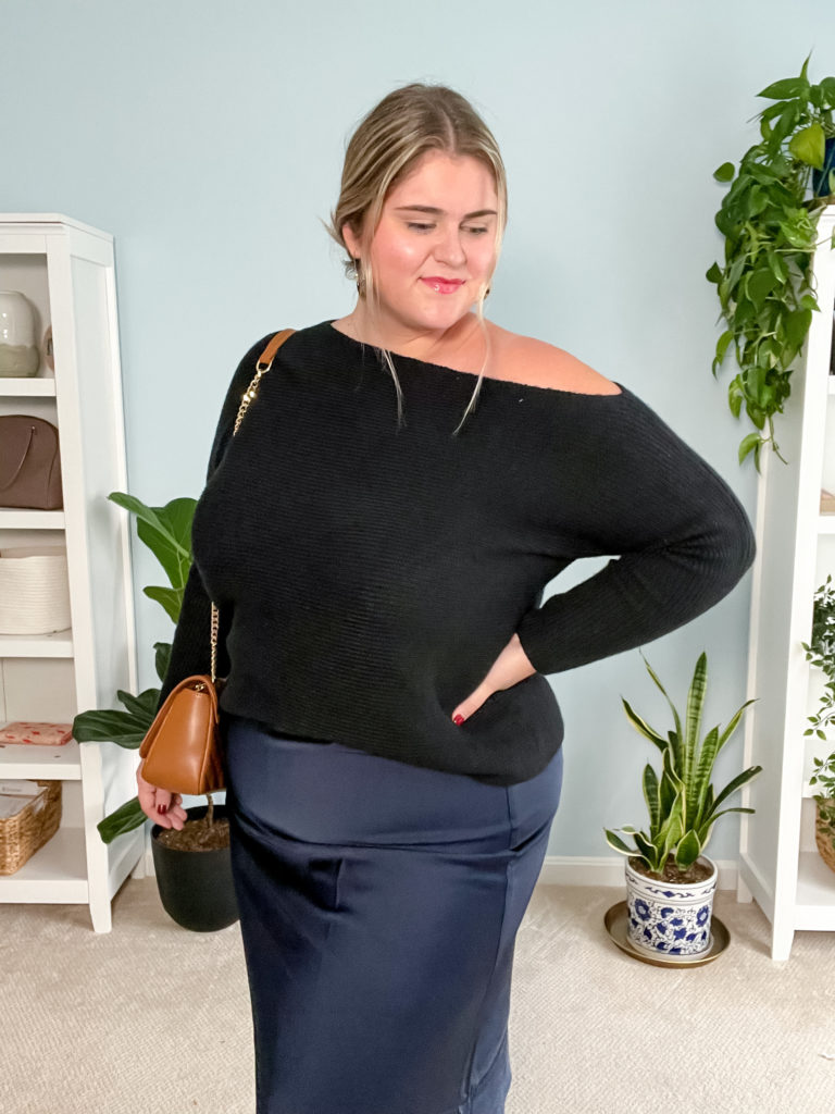A happy plus-size woman is wearing a black sweater and navy slip skirt while posing in her bedroom. 