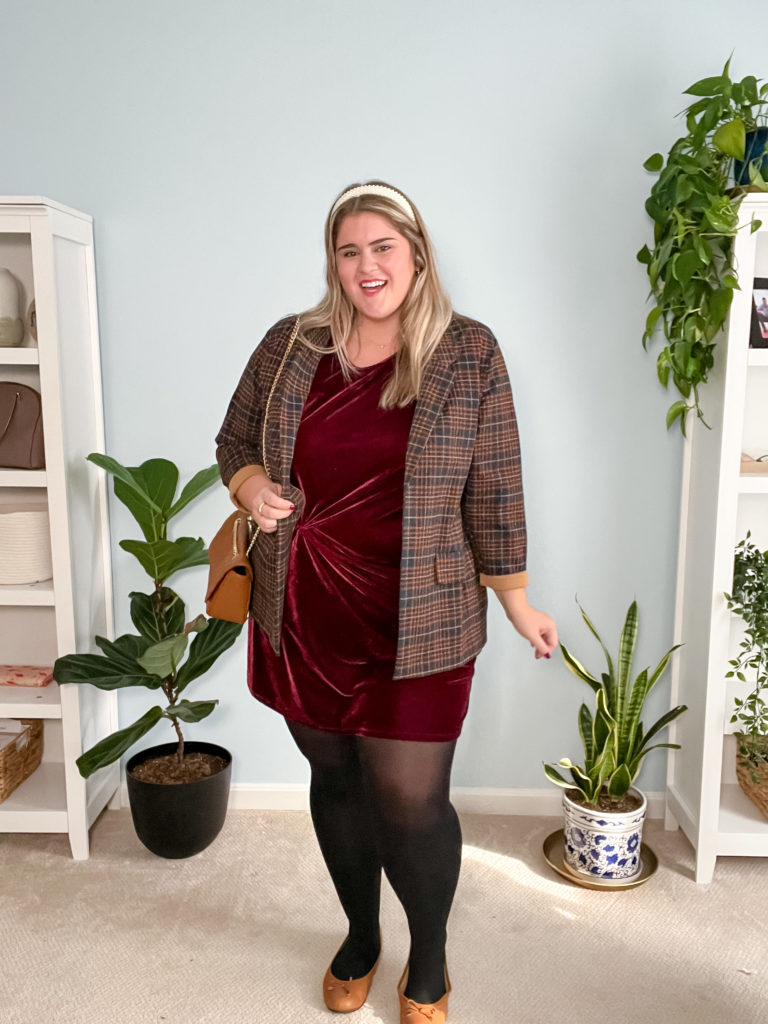 A happy Caucasian woman wearing a burgundy velvet dress and plaid blazer as an example of jackets that go with dresses.