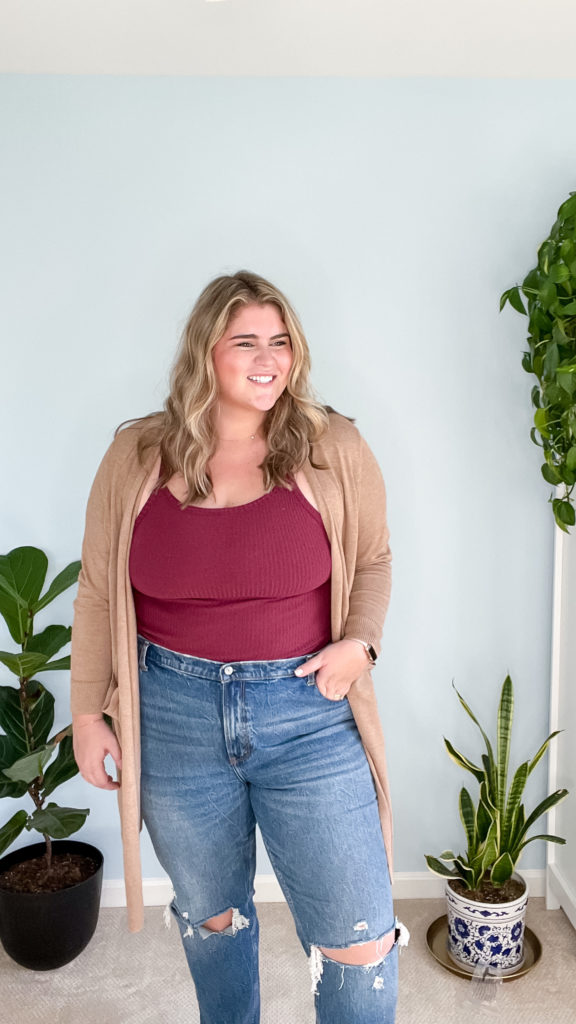 A happy plus-size blonde woman is posing for a picture in her room wearing a burgundy tank top, tan cardigan and jeans. 