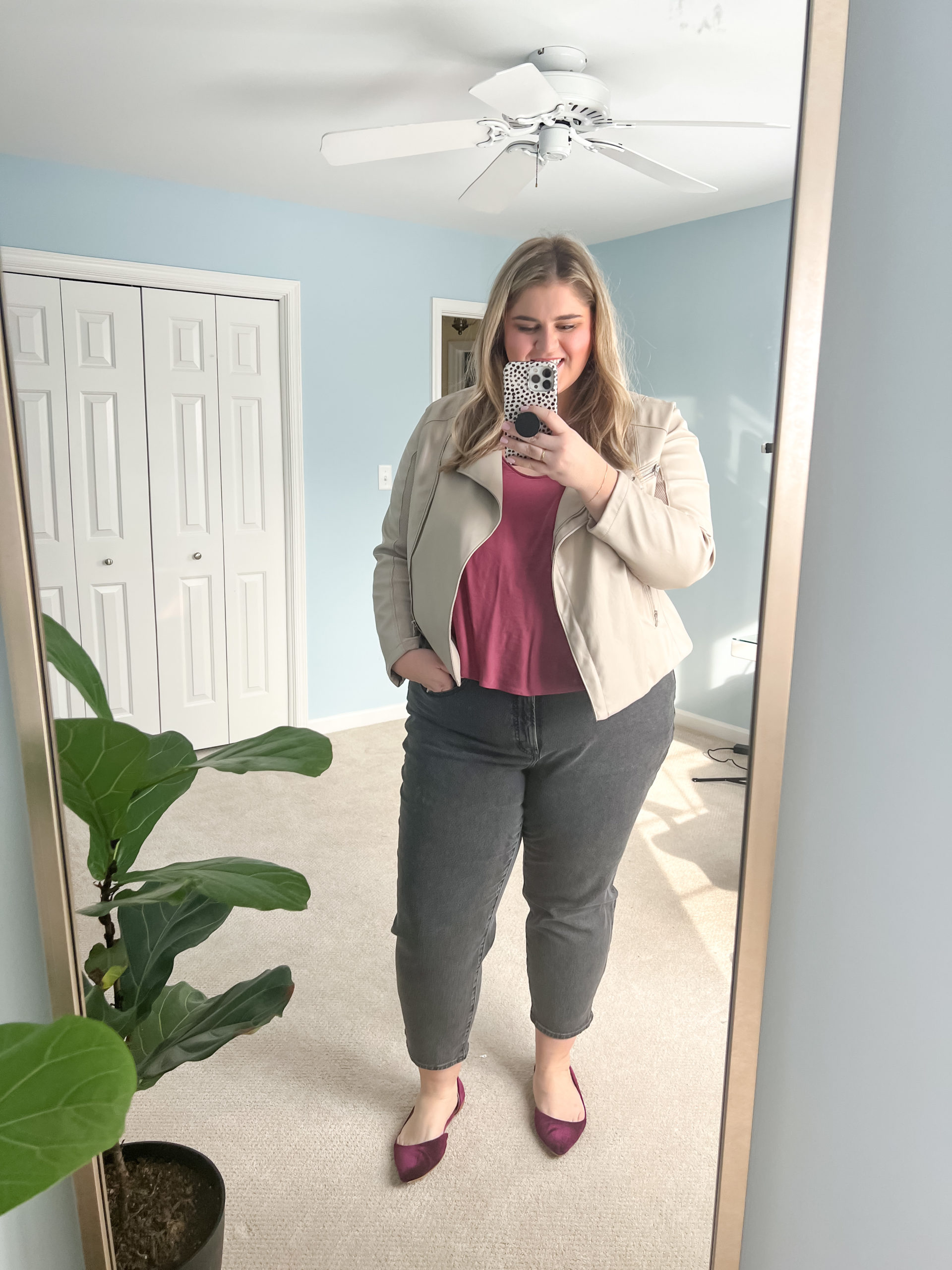 a plus size blonde woman taking a mirror selfie in her bedroom wearing black straight leg jeans, a burgundy top and a moto jacket with velvet flats