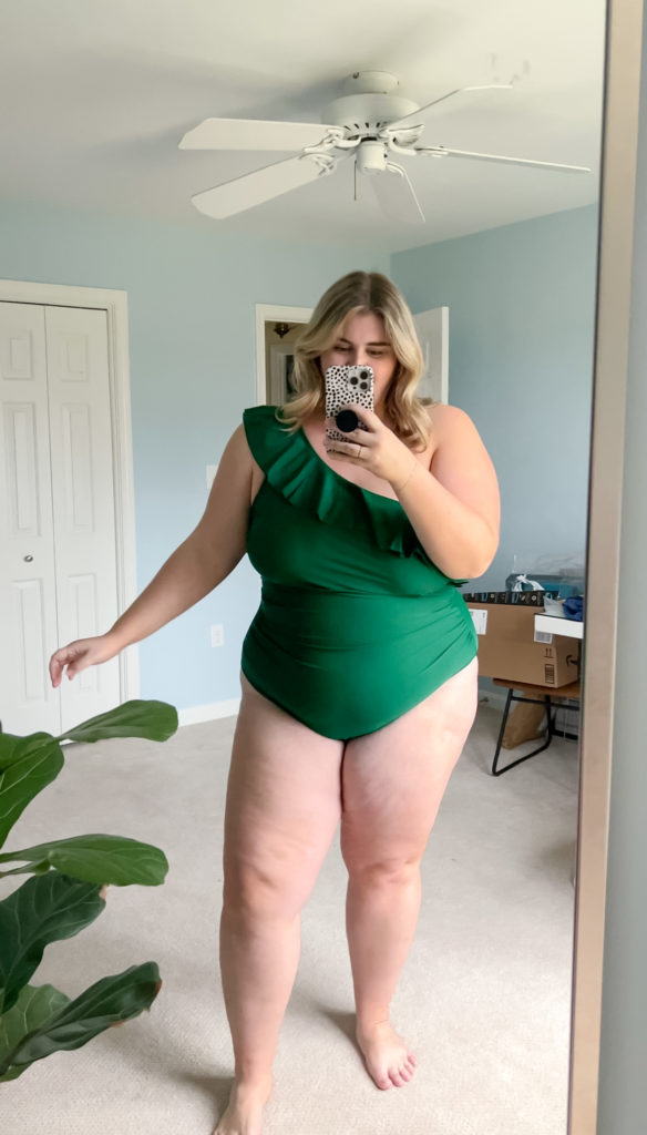 A Caucasian blonde woman wearing a green Amazon plus size one-piece bathing suit in her bedroom while taking a mirror selfie. 