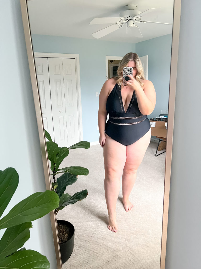 A plus size blonde woman wearing a black one piece swimsuit in her bedroom while taking a mirror seflie.