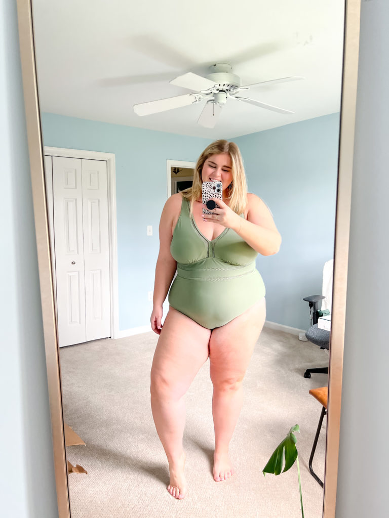 A smiling blonde woman modeling an Amazon plus size swimsuit in her bedroom that is sage green.