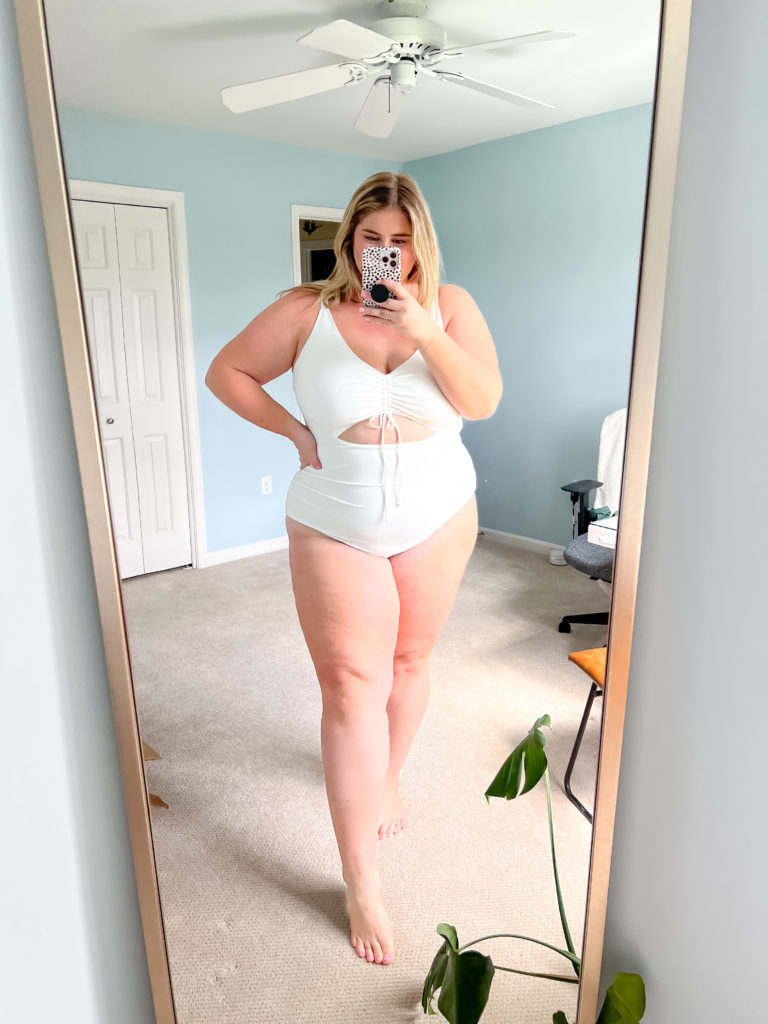 A happy Caucasian woman taking a mirror selfie while modeling a white one piece bathing suit from Amazon. 