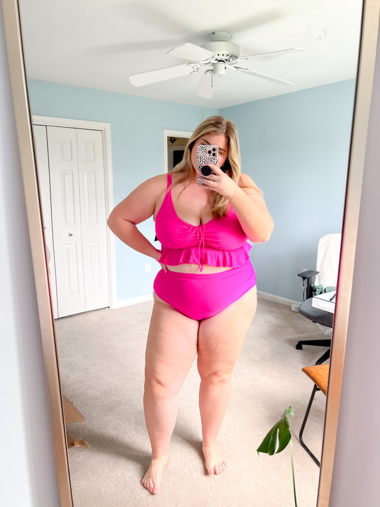 A plus size blonde woman is taking a mirror selfie in her bedroom while wearing a hot pink bikini. 