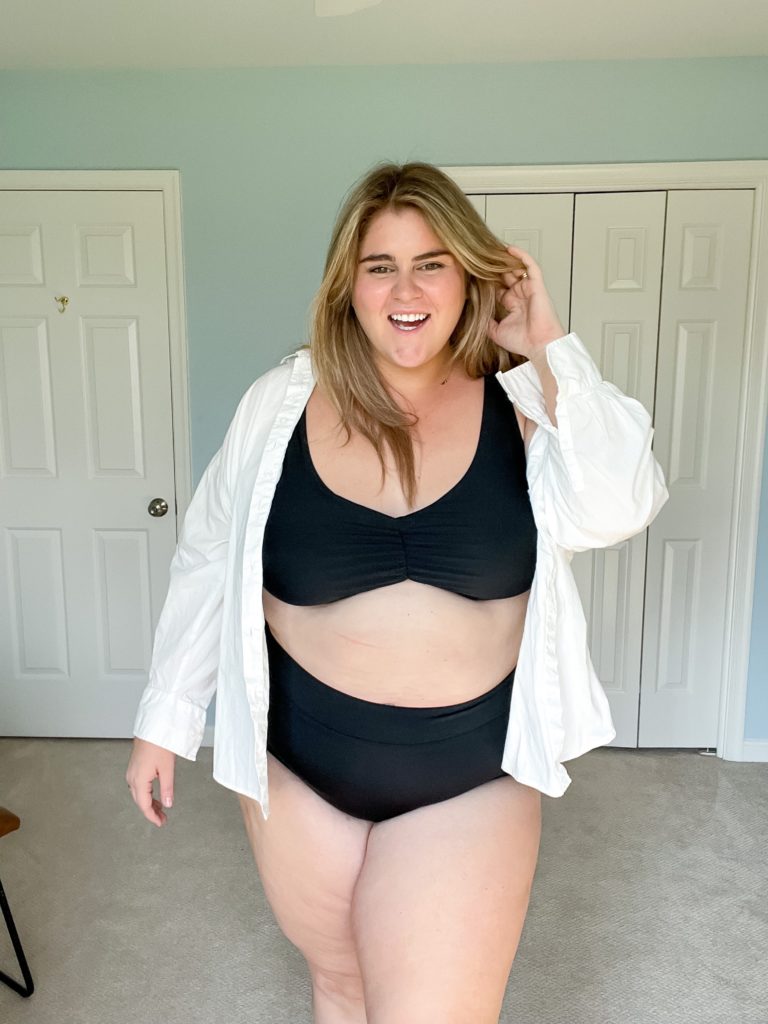 A happy Caucasian woman is wearing a plus-size black bikini and white shirt in her bedroom, smiling for the picture. 