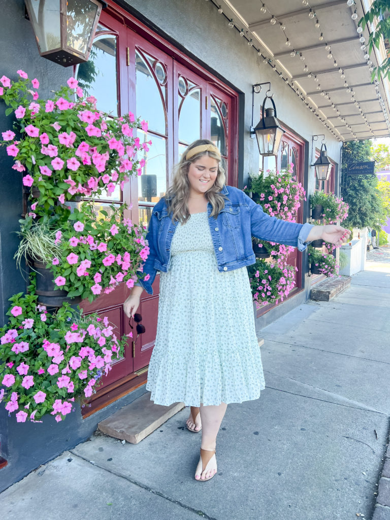 A blonde woman wearing a floral dress and denim jacket standing in front of a shop.