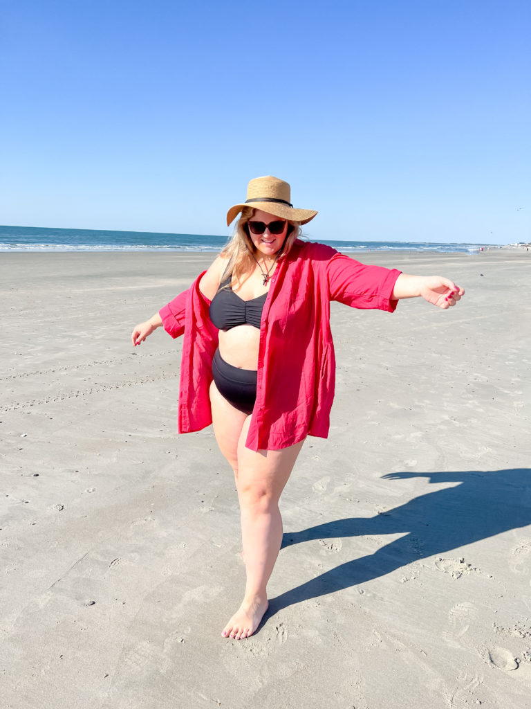 A smiling blonde woman is wearing a black bikini and red swim cover-up while standing on the beach with her arms out. 