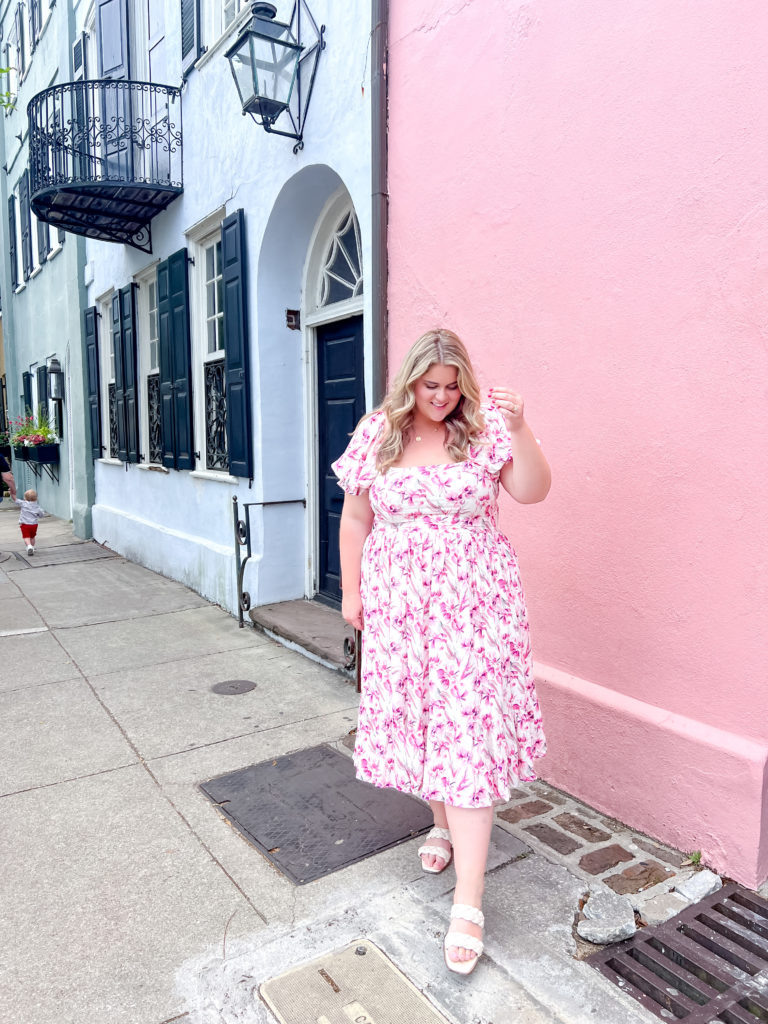 A happy Caucasian woman is posing for a picture in front of the pink Charleston Row house wearing a plus-size Jessa Kae floral dress. 