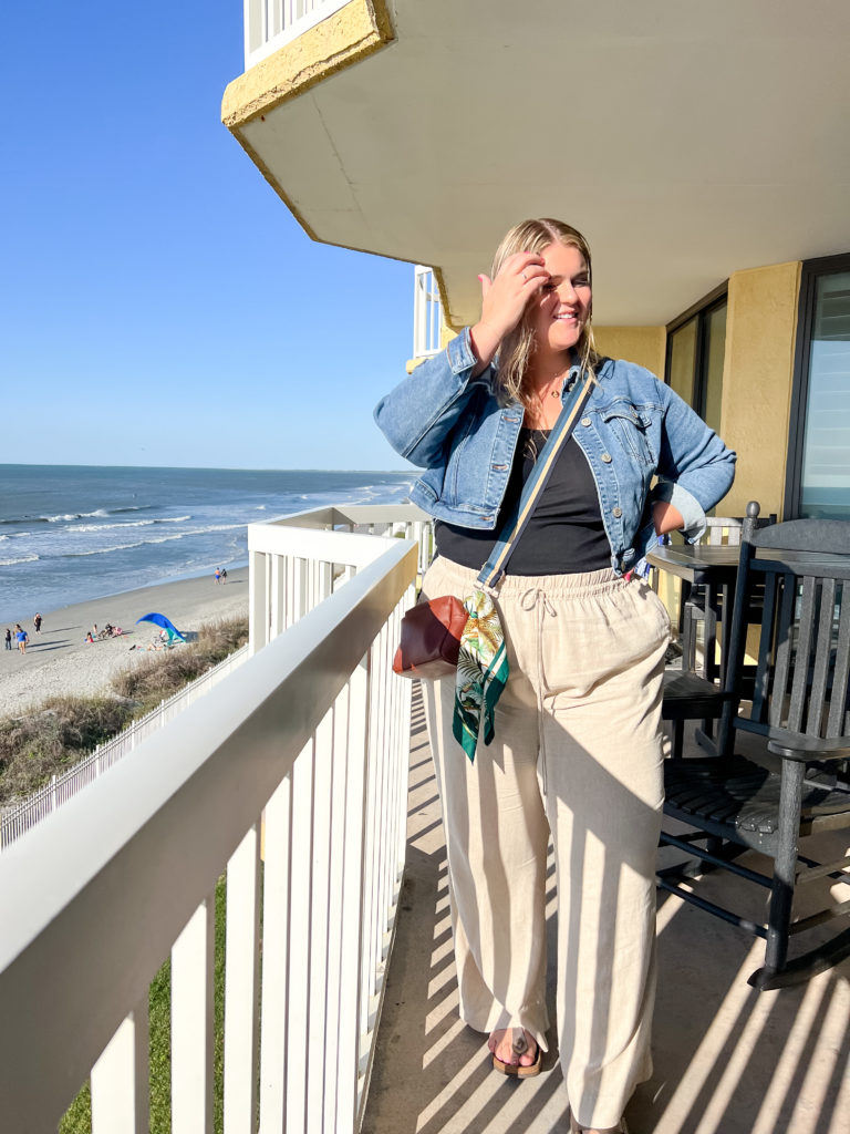 A blonde woman standing on a balcony overlooking the beach, wearing a vacation outfit in plus size of linen pants, black tank and denim jacket. 