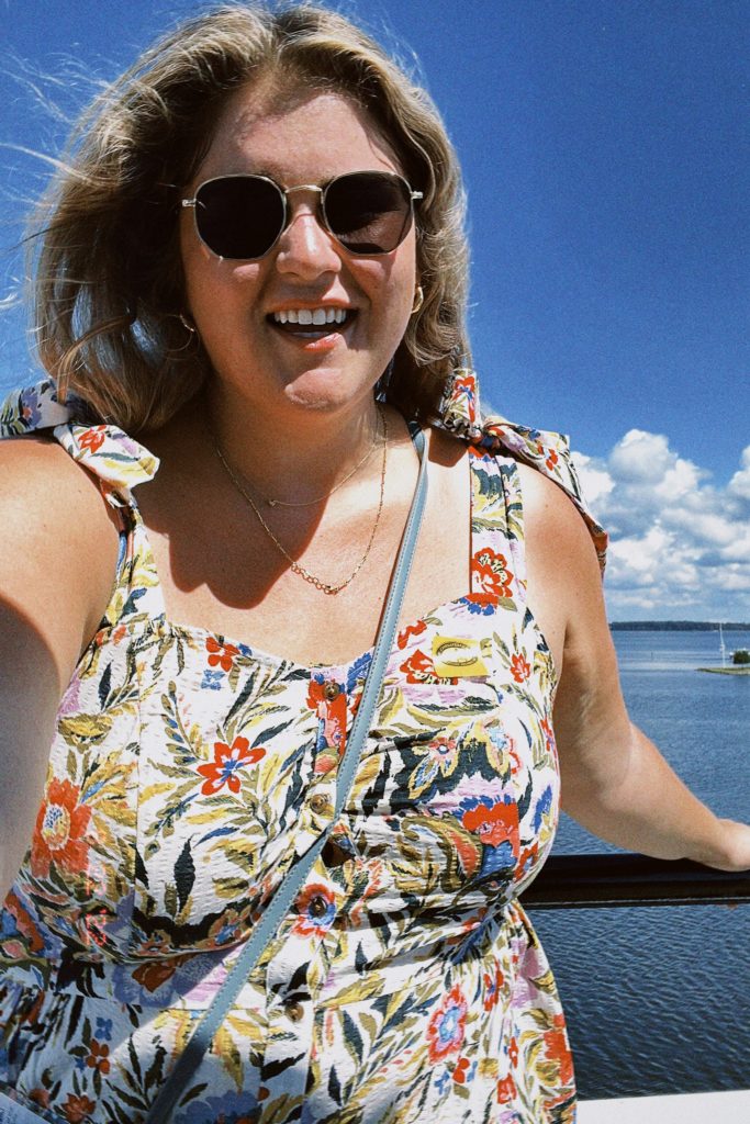 A smiling Caucasian woman is taking a selfie on a boat while wearing a plus size floral dress. 