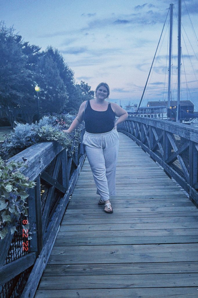 A blonde woman is wearing a plus size vacation outfit of linen pants and a black tank top. 
