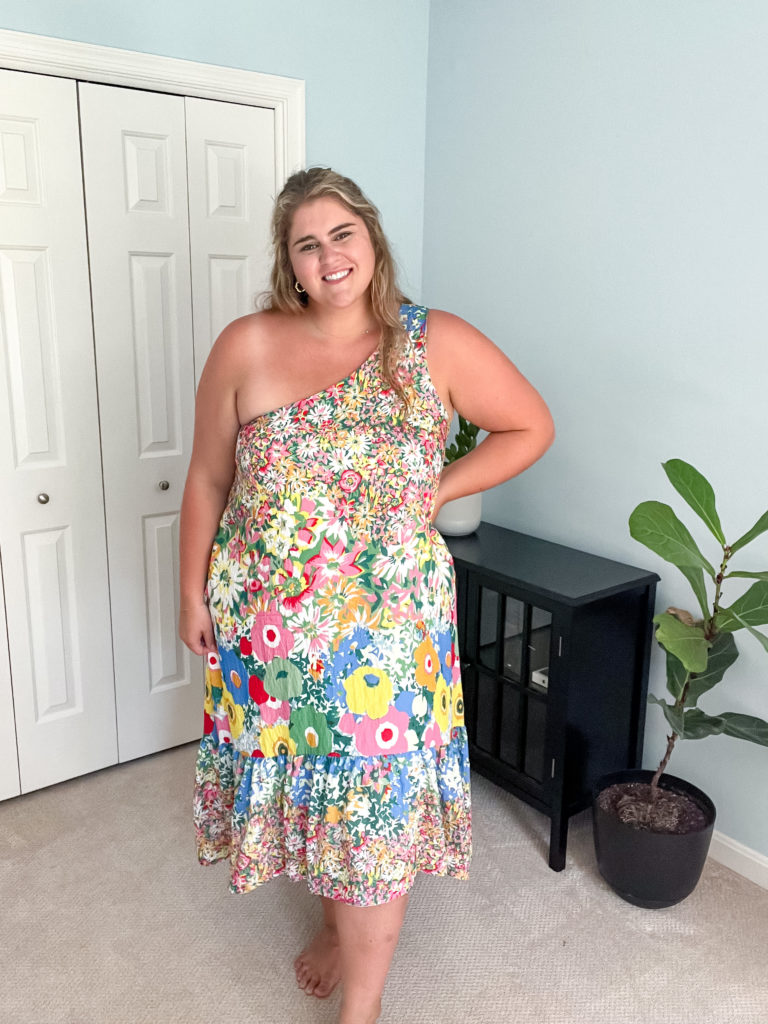 A smiling Caucasian woman is posing for a picture in her bedroom while wearing a floral midi dress. 