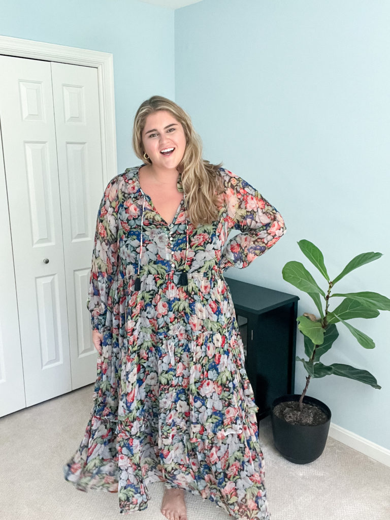 A happy, blonde plus size woman wearing a multicolor floral maxi dress in her bedroom. 