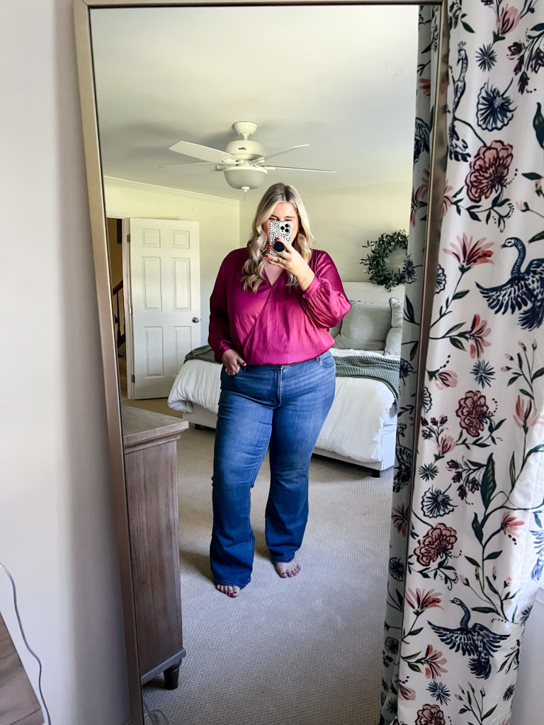 A Caucasian woman is taking a mirror selfie wearing a raspberry colored top and dark denim jeans. 