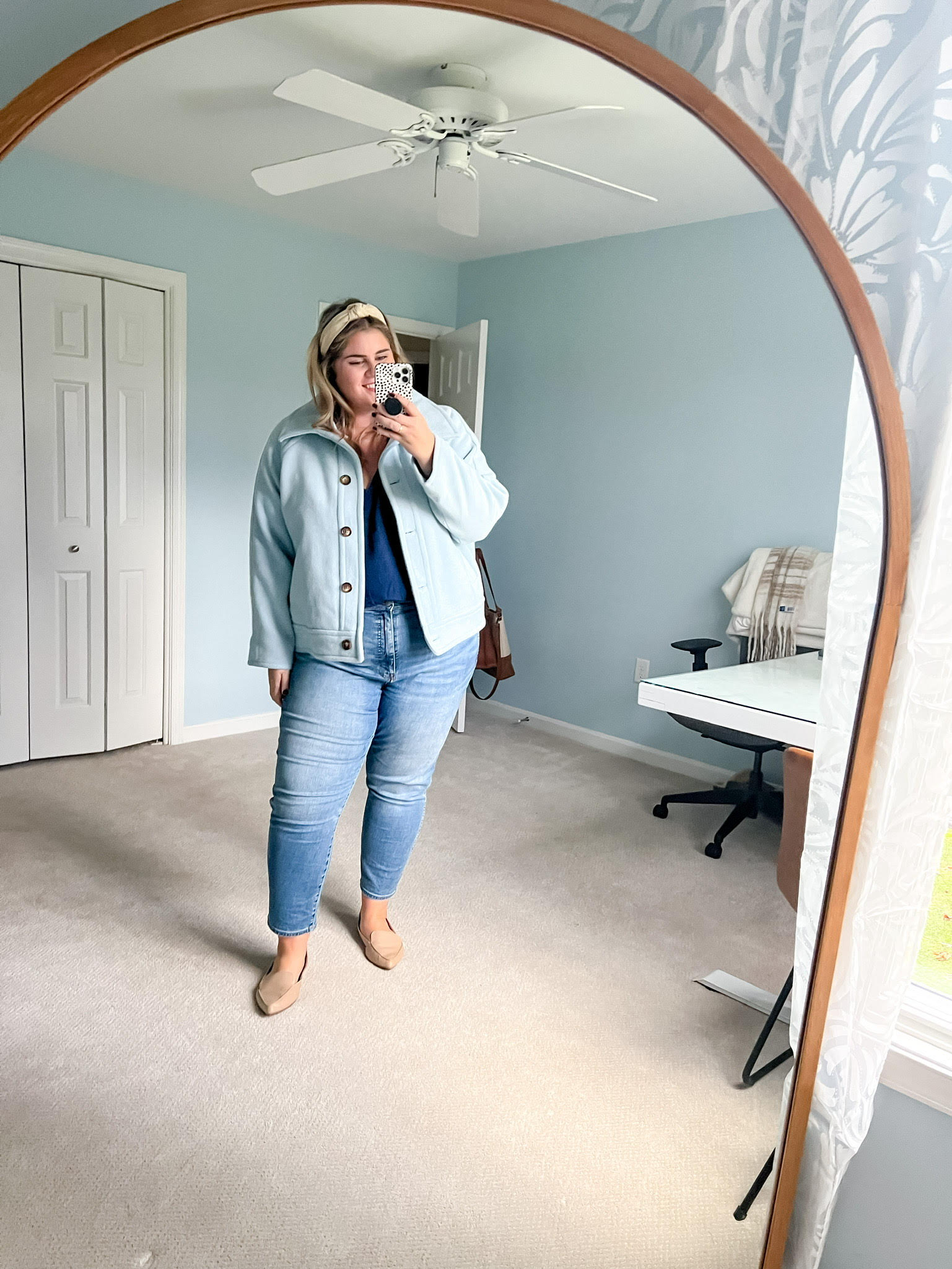 a happy blonde woman taking a mirror selfie wearing a blue coat, teal top, jeans and tan loafers