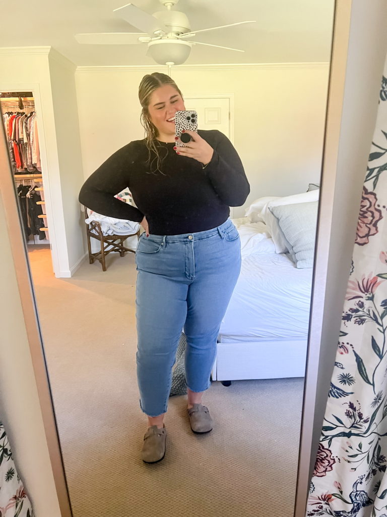 a happy blonde woman taking a mirror selfie wearing a black shirt, straight leg jeans and birks