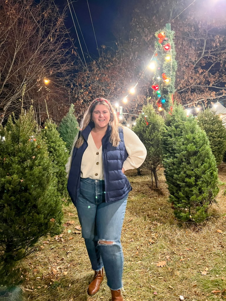 A happy Caucasian woman is posing at a Christmas tree lot wearing a plus-size brand Lands' End outfit of jeans, boots, sweater and puffer vest. 