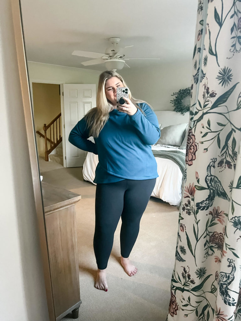 A smiling Causcasian woman is taking a mirror selfie wearing black leggings and a blue top.