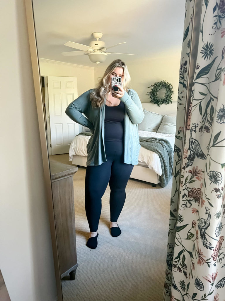 A blonde woman is taking a mirror selfie in her bedroom wearing leggings from her favorite plus-size brand Athleta.