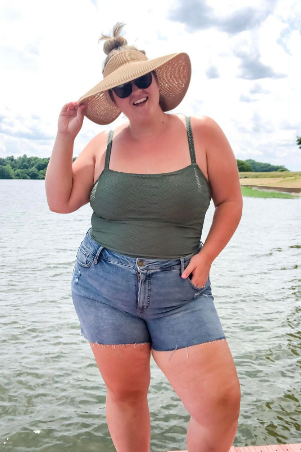 A plus size Caucasian woman wearing a green bathing suit, denim shorts and straw hat is posing for a picture by the lake. 