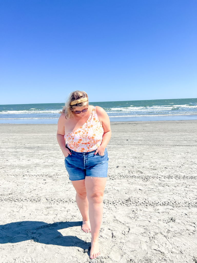 A plus size Caucasian woman is wearing a floral bathing suit and denim shorts on the beach. 