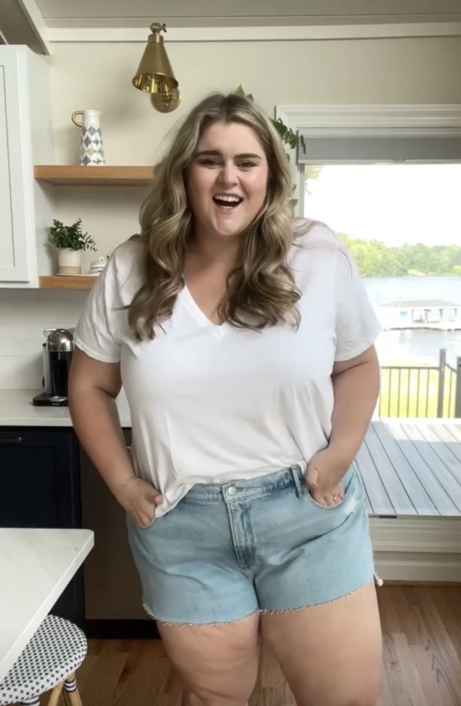 A happy blonde woman wearing a white tee and denim shorts in her kitchen.
