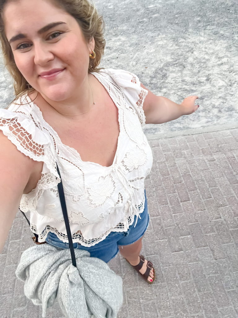A smiling blonde woman is taking a selfie on the sidewalk wearing a vacation outfit in plus size.