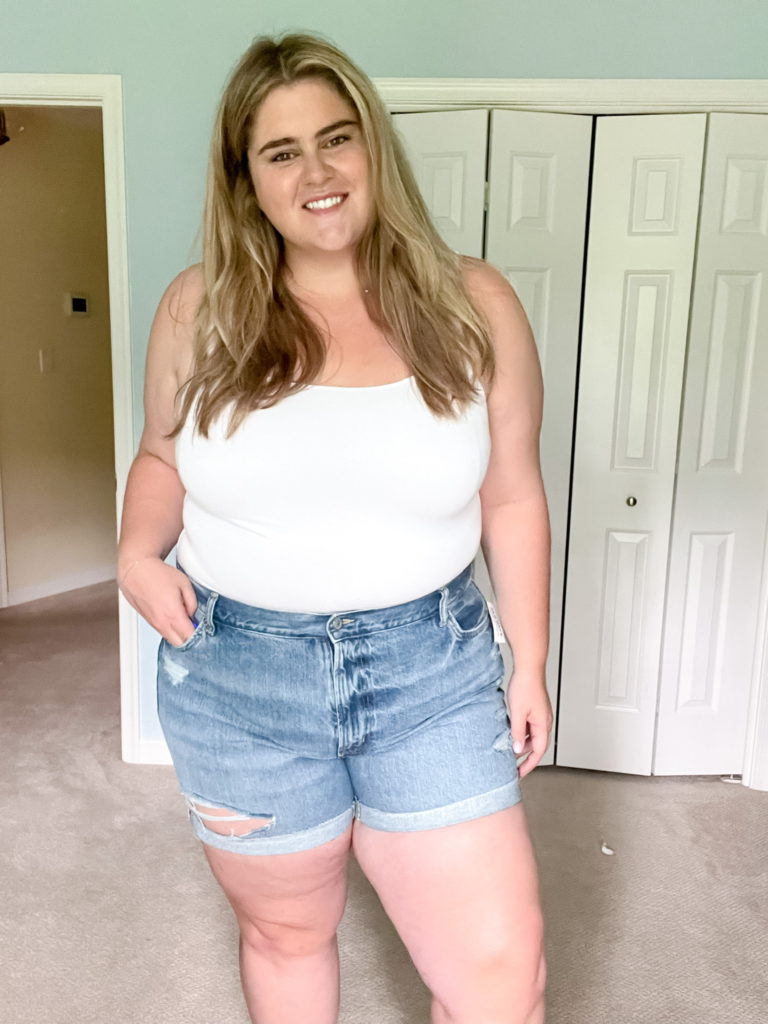 A smiling Caucasian woman is posing for a picture wearing a white tank top and denim shorts from a beloved plus-size brand Old Navy. 