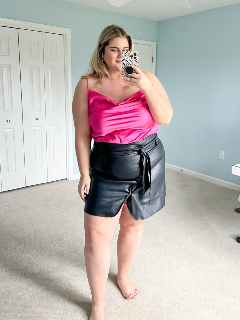 A Caucasian woman is wearing a hot pink top and black mini skirt while taking a mirror selfie in her bedroom. 