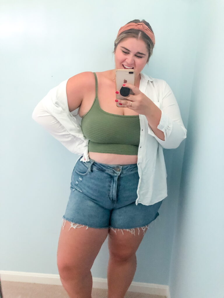 A smiling Caucasian woman is taking a mirror selfie while wearing a green bikini top, denim shorts and a white button down. 