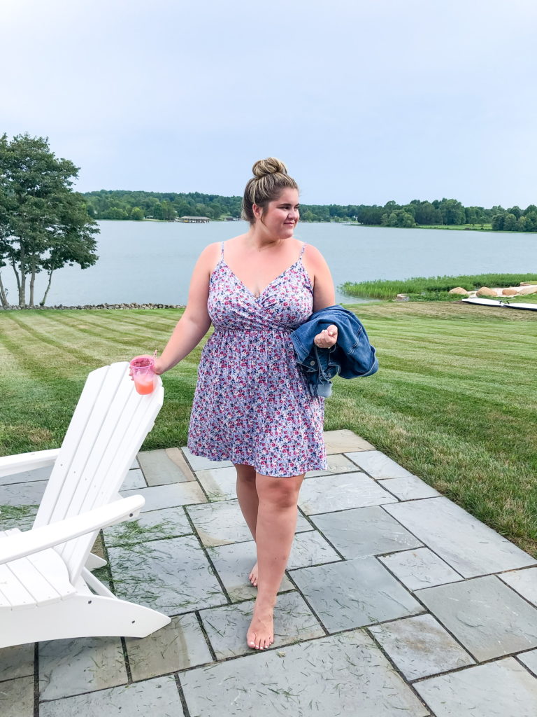 A blonde woman wearing a short floral dress as a plus size vacation outfit at a lake house.