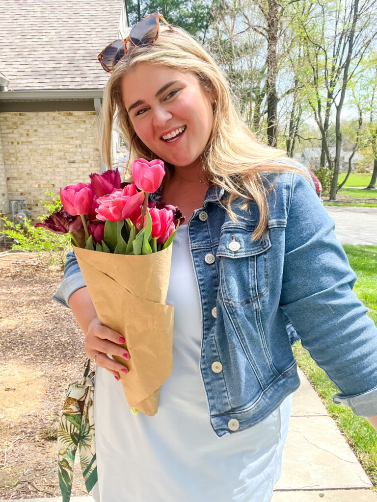 A happy Caucasian woman is holding a bouquet of tulips and denim jacket while standing outside of her house. 