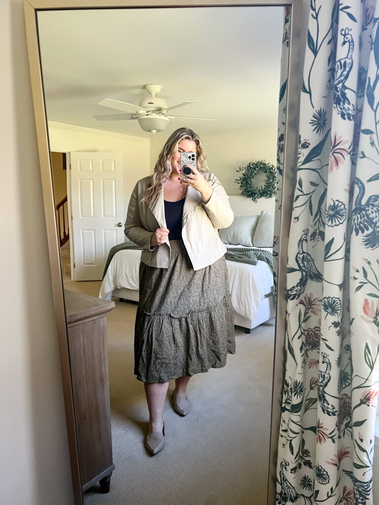 A plus size woman is taking a mirror selfie while wearing a midi skirt, black tank top and cream moto jacket. 