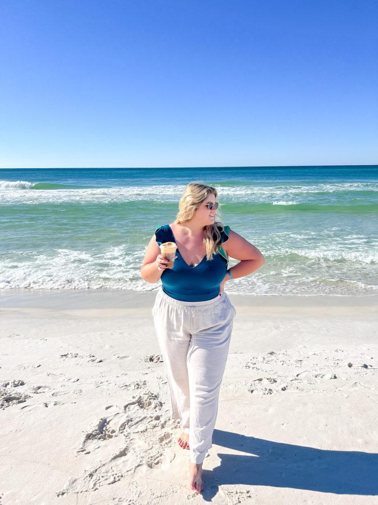 A Caucasian woman standing on the beach wearing a bath suit and linen pants as a vacation outfit in plus size.