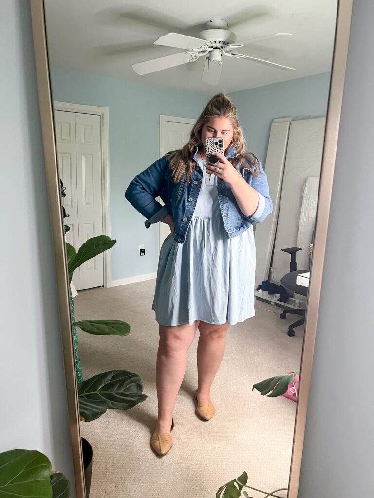 A Caucasian woman is taking a mirror selfie while modeling a jacket that goes with dresses. 