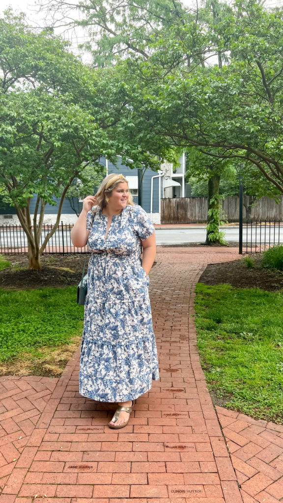 A smiling blonde woman wearing a white and blue floral maxi dress as a vacation outfit in plus size, standing on a path in a park. 