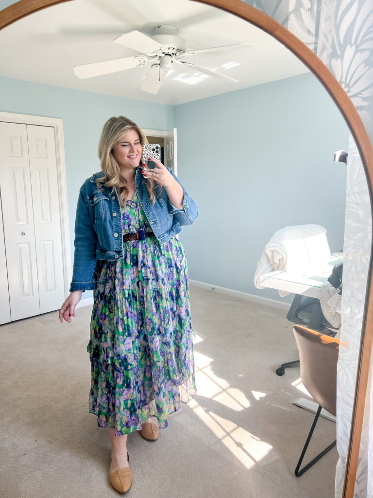 A smiling plus-size woman is taking a mirror selfie wearing a floral Anthrolpolgie dress and denim jacket. 